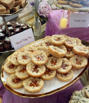 Golden apricot baby pies with a buttery shortbread base and brown sugar streusel topping, displayed on a white and gold tray at a wedding dessert table. Styled with Parisian-inspired florals and soft fabrics at the historic Dr. Phillips House in Orlando, Florida. A signature treat by The Cookie Jar Orlando.