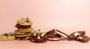 A colorful variety of gourmet cookies including chocolate chip, red velvet, funfetti, peppermint, sandwich cookies with buttercream filling, and gooey marshmallow-stuffed chocolate cookies, arranged against a soft pink backdrop. An ideal selection from The Cookie Jar Orlando for wedding dessert tables, party trays, and custom cookie favors.
