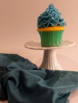 A vanilla cupcake topped with sky-blue buttercream swirls and delicate sprinkle accents, displayed in a matching teal wrapper on a white pedestal. Draped with soft teal fabric for an elegant presentation, this cupcake is ideal for baby showers, birthday celebrations, or winter-themed parties from The Cookie Jar Orlando.