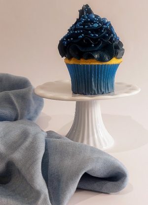 Vanilla cupcake topped with rich blue buttercream frosting, decorated with assorted blue sprinkles and pearls. Presented on a white cake stand next to soft blue fabric, this cupcake from The Cookie Jar Orlando is perfect for baby showers, winter-themed events, and upscale corporate gatherings.