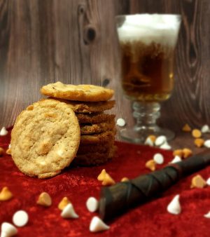 Golden butterbeer cookies with white chocolate and butterscotch chips, stacked on red velvet with a glass of frothy cream soda and a wand in the background. A magical treat by The Cookie Jar Orlando, perfect for Harry Potter-themed parties, birthdays, and whimsical dessert tables.