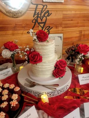Two-tier buttercream wedding cake with ridged frosting, decorated with vibrant red roses and baby’s breath, topped with a custom “Tyla ♥︎ MZ” topper. Displayed on a red and white dessert table at Club Lake Venue in Apopka, Florida. Created by The Cookie Jar Orlando.