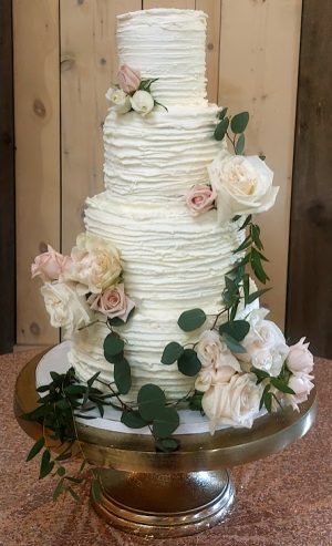 Four-tier classic white wedding cake with sweet cream buttercream and ruffle-textured frosting, decorated with blush roses and eucalyptus greenery. Displayed on a gold cake stand at The Barn at Hidden Oaks Farm wedding venue in Lake Mary, Florida. Created by The Cookie Jar Orlando.
