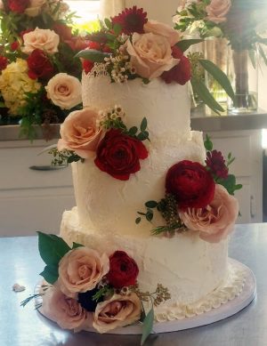 Three-tier buttercream wedding cake with delicate lace fondant detailing, adorned with fresh blush and deep red roses. Designed for a romantic wedding at the historic Howey Mansion in Central Florida by The Cookie Jar Orlando.