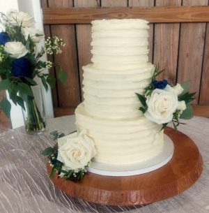 Three-tier buttercream wedding cake with ridged frosting texture, decorated with white roses, greenery, and touches of royal blue. Displayed on a wood cake stand for a romantic wedding at Pegasus Manor near Orlando, Florida. Created by The Cookie Jar Orlando.