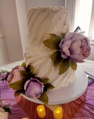 Two-tier buttercream wedding cake with twisted texture and large lavender peony florals, displayed on a mirrored base with candlelight at Historic Venue 1902 in Sanford, Florida. Designed by The Cookie Jar Orlando for a vintage-inspired celebration.