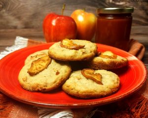 Soft caramel apple cookies topped with baked apple slices, served on a bright red plate with fresh apples and a jar of caramel in the background. A seasonal favorite by The Cookie Jar Orlando, perfect for fall weddings, dessert tables, and cozy corporate gatherings.