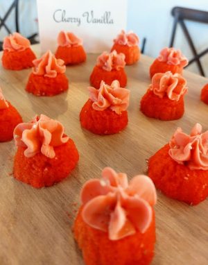 Mini cherry vanilla bundt cakes with pink buttercream swirls, arranged on a wood tray at a rustic wedding dessert table at The Wedding Barn in Oviedo, Florida. A colorful and festive treat by The Cookie Jar Orlando — ideal for weddings, birthday parties, sweet sixteens, and summer celebrations.