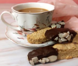 Handmade almond biscotti cookies dipped in dark chocolate and topped with sliced almonds, styled with a floral teacup and blush pink background. Ideal for weddings, bridal showers, tea parties, or corporate gifting in Central Florida. Created by The Cookie Jar Orlando.