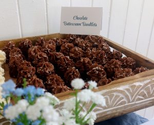 Mini chocolate bundt cakes topped with rich chocolate fudge buttercream and white sugar pearls, displayed in a carved wooden tray with florals. Served on a vintage wedding dessert table at Historic Venue 1902 in Sanford, Florida. A signature sweet by The Cookie Jar Orlando, perfect for weddings, showers, and guest favors.