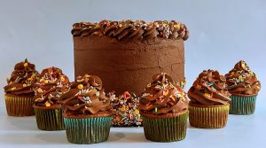 A chocolate frosted celebration cake surrounded by matching cupcakes, all topped with colorful sprinkles and piped chocolate buttercream. Perfect for birthdays, parties, baby showers, and graduations, this cheerful set from The Cookie Jar Orlando brings fun and flavor to any festive occasion.