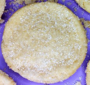 A golden, round sugar cookie topped with a sparkling layer of coarse sugar crystals, set on a vibrant purple background. A timeless treat by The Cookie Jar Orlando, perfect for weddings, party trays, corporate events, and edible favors.