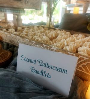 Mini coconut bundt cakes topped with creamy buttercream swirls, displayed in a carved wooden tray at a tropical-inspired wedding dessert table at Paradise Cove in Orlando. A signature treat by The Cookie Jar Orlando, perfect for weddings, summer parties, and beach-themed celebrations.