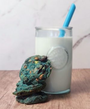 Vibrant blue Cookie Monster cookies with chocolate chips and candy bits, stacked beside a glass of milk with a blue straw. A whimsical treat by The Cookie Jar Orlando, perfect for children’s parties, school celebrations, and fun dessert tables.