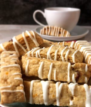 Cranberry orange biscotti cookies drizzled with white chocolate, stacked artfully beside a teacup for a soft, elegant display. A fresh and flavorful option for weddings, bridal showers, or corporate gifting in Central Florida. Crafted by The Cookie Jar Orlando.