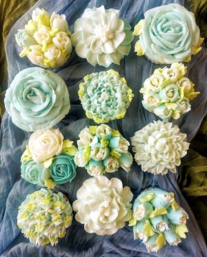 A dozen cupcakes decorated with intricate pastel buttercream flowers in soft shades of blue, green, and white, arranged on draped fabric – ideal for weddings, bridal showers, baby showers, and Mother's Day events.