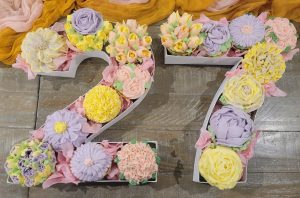 A stunning arrangement of buttercream flower cupcakes in the shape of the number 27, featuring soft pastel shades of yellow, lavender, and pink—perfect for birthdays, bridal showers, or garden-themed celebrations.