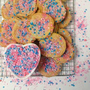Brightly colored funfetti birthday cake cookies topped with pink, blue, and white sprinkles, arranged on a cooling rack with a heart-shaped dish of matching sprinkles. A festive and fun dessert option from The Cookie Jar Orlando, ideal for birthday parties, kids' celebrations, and event favors.