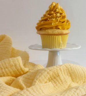 A single vanilla cupcake topped with bright yellow buttercream swirls and golden star sprinkles, elegantly displayed on a white pedestal against a cozy, soft yellow fabric. Perfect for baby showers, graduations, and joyful celebrations. A cheerful, handcrafted treat by The Cookie Jar Orlando.
