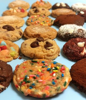 An assortment of colorful gourmet cookies including chocolate chip, red velvet, sugar, funfetti, double chocolate, and cookies with candy pieces, arranged in neat rows on a soft blue background. A versatile selection from The Cookie Jar Orlando, perfect for corporate events, wedding dessert tables, party trays, and celebration favors.