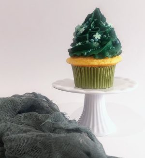 Vanilla cupcake topped with deep green buttercream frosting and adorned with dainty sugar flowers and pearl sprinkles. Displayed on a white cake pedestal beside textured green fabric, this cupcake is ideal for holiday dessert tables, St. Patrick’s Day parties, and whimsical garden celebrations by The Cookie Jar Orlando.