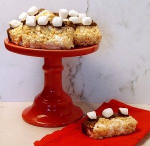 Marshmallow-topped Rice Krispy treats dipped in chocolate, displayed on a vibrant red cake stand with a matching napkin—ideal for birthday parties, kids events, and sweet celebrations in Central Florida.