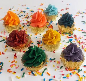 Assorted mini vanilla cupcakes topped with vibrant buttercream swirls in gold foil wrappers, arranged on a white surface with rainbow sprinkles. These festive bite-sized treats from The Cookie Jar Orlando are ideal for birthday parties, wedding dessert tables, showers, and corporate celebrations.