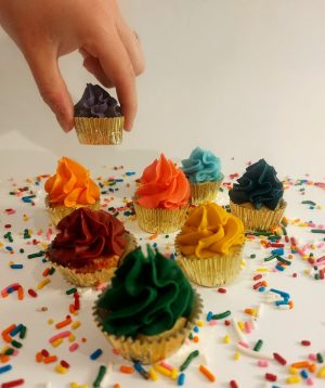 A hand reaching for a mini cupcake with purple buttercream frosting, surrounded by other colorful mini cupcakes in gold foil wrappers. Set on a white surface with rainbow sprinkles, these festive treats from The Cookie Jar Orlando are perfect for kids’ birthday parties and celebrations.