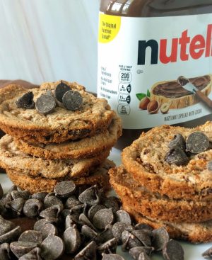 Stacked Nutella chocolate chip cookies surrounded by rich chocolate chips and a Nutella jar in the background. A chewy, decadent cookie by The Cookie Jar Orlando, perfect for wedding dessert tables, corporate events, birthday parties, and celebration favors.