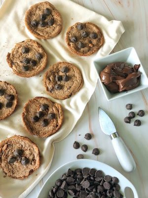 Nutella chocolate cookies arranged on a cream cloth with bowls of chocolate chips and Nutella spread. A luxurious cookie option from The Cookie Jar Orlando, ideal for wedding dessert tables, event trays, party platters, and upscale cookie favors.