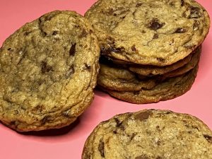 Golden-brown old-fashioned chocolate chip cookies with crisp edges and gooey centers, stacked on a vibrant pink background. A beloved classic from The Cookie Jar Orlando, ideal for wedding dessert tables, party trays, and client gift favors.
