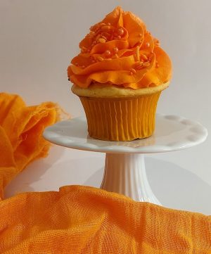 A festive vanilla cupcake crowned with bright orange buttercream and orange sprinkles, sitting on a white pedestal surrounded by vivid orange fabric. A cheerful treat by The Cookie Jar Orlando, ideal for birthdays, autumn parties, Halloween dessert tables, and fall-themed celebrations.