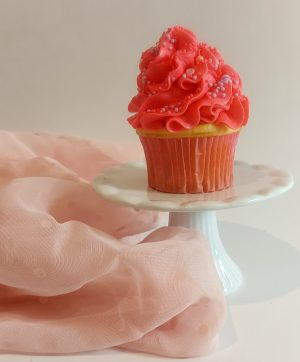 A charming vanilla cupcake swirled with bright pink buttercream frosting and decorated with tiny pastel pink and white sprinkles. Displayed on a white cake stand and surrounded by soft blush fabric, this cupcake is perfect for baby showers, bridal celebrations, and Valentine’s Day desserts from The Cookie Jar Orlando.