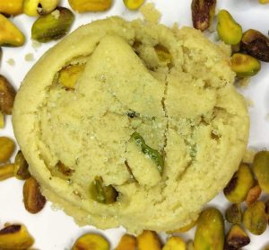 Close-up of a soft pistachio sugar cookie sprinkled with pale green sanding sugar and surrounded by whole pistachios. A unique and elegant choice for wedding dessert tables, cookie favors, and corporate or holiday event trays by The Cookie Jar Orlando.