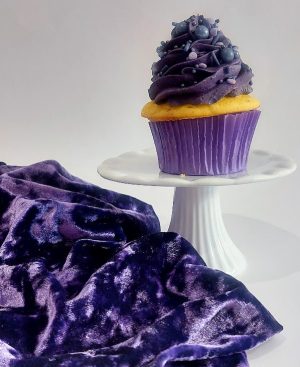 A vibrant cupcake with deep purple buttercream swirls, topped with pearl sprinkles in various shades of purple. Presented in a matching purple wrapper on a white pedestal, with plush purple velvet fabric adding a rich, festive backdrop. Ideal for Halloween dessert tables, birthday parties, or kids’ celebrations from The Cookie Jar Orlando.