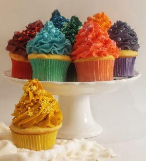 A vibrant assortment of vanilla cupcakes topped with brightly colored buttercream swirls, each in a different shade of the rainbow and decorated with themed sprinkles. Displayed on a white pedestal cake stand, ideal for birthdays, celebrations, and party dessert tables. A festive treat by The Cookie Jar Orlando.