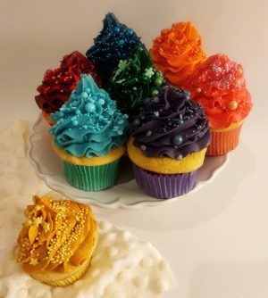 A cheerful display of vanilla cupcakes topped with vivid buttercream swirls in rainbow colors, adorned with decorative sprinkles. Arranged on a white ruffled platter with one cupcake styled separately in the foreground. Ideal for birthdays, parties, and festive events, these cupcakes are a playful signature from The Cookie Jar Orlando.