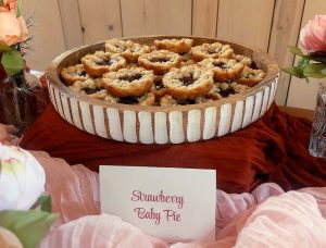 Strawberry baby pies with French shortbread crust and brown sugar streusel filling, displayed in a rustic wood tray surrounded by soft florals and pink linen. Styled for a wedding at The Barn at Hidden Oaks Farm in Lake Mary, Florida. A romantic dessert option by The Cookie Jar Orlando.