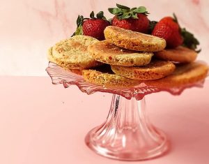 Golden strawberry shortcake cookies displayed on a pink glass stand with fresh strawberries, perfect for weddings and summer parties.