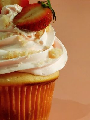 Close-up of a vanilla cupcake topped with pink strawberry buttercream, fresh strawberry halves, and cake crumbs. A charming dessert option for bridal showers, baby showers, and Mother’s Day celebrations.