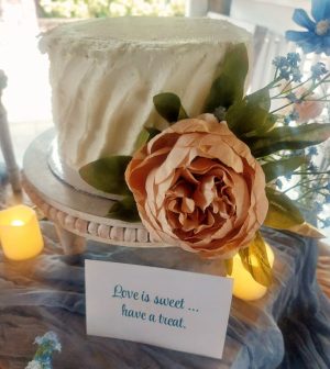 Small sweetheart buttercream wedding cake with textured sides, adorned with a peach garden rose and greenery. Displayed on a vintage-style white stand with soft candlelight and a sign that reads “Love is sweet... have a treat.” Created for a wedding at Paradise Cove in Orlando, Florida by The Cookie Jar Orlando.