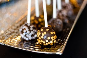 Glamorous chocolate truffle cake pops decorated with gold and silver confetti sprinkles, elegantly arranged on a metallic tray. Styled for a modern wedding dessert table at New York Beer Project in Orlando, these rich chocolate-coated treats from The Cookie Jar Orlando are perfect for upscale weddings, sweet sixteens, and luxury parties.