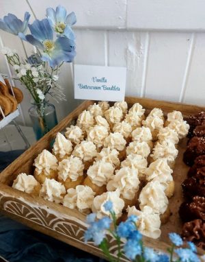Mini vanilla bundt cakes topped with swirls of creamy buttercream, arranged in a carved wood tray beside fresh blue florals. Displayed as part of an elegant wedding dessert table at Historic Venue 1902 in Sanford, Florida. A handcrafted treat by The Cookie Jar Orlando, perfect for weddings, bridal showers, birthdays, and baby celebrations.