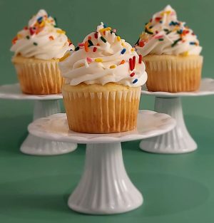 Three vanilla cupcakes topped with white buttercream and rainbow sprinkles, each displayed on individual mini white cake stands—perfect for birthdays, celebrations, and party dessert tables.